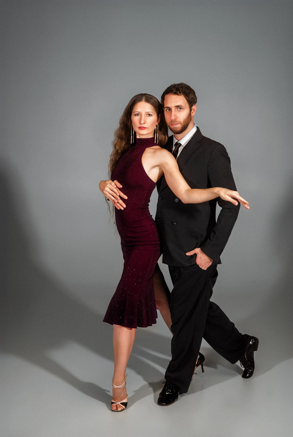 A full-body studio portrait of a dance couple posing against a plain grey background. The woman, standing in front, wears a sleeveless burgundy dress and dance heels, extending her left arm outward while resting her right hand on her torso. The man stands slightly behind her in a black double-breasted suit with one hand in his pocket, and both look directly at the camera with serious expressions.