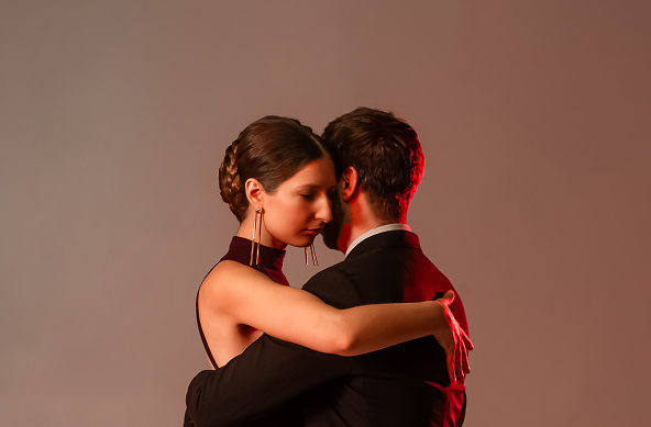 A close-up profile shot of the couple in a romantic tango embrace, illuminated by dramatic red lighting. The woman has her eyes closed and her forehead resting gently against the man's cheek, showcasing her braided hair and long gold earrings. The man is viewed from the back, wearing a black suit, holding her in a close dance frame.