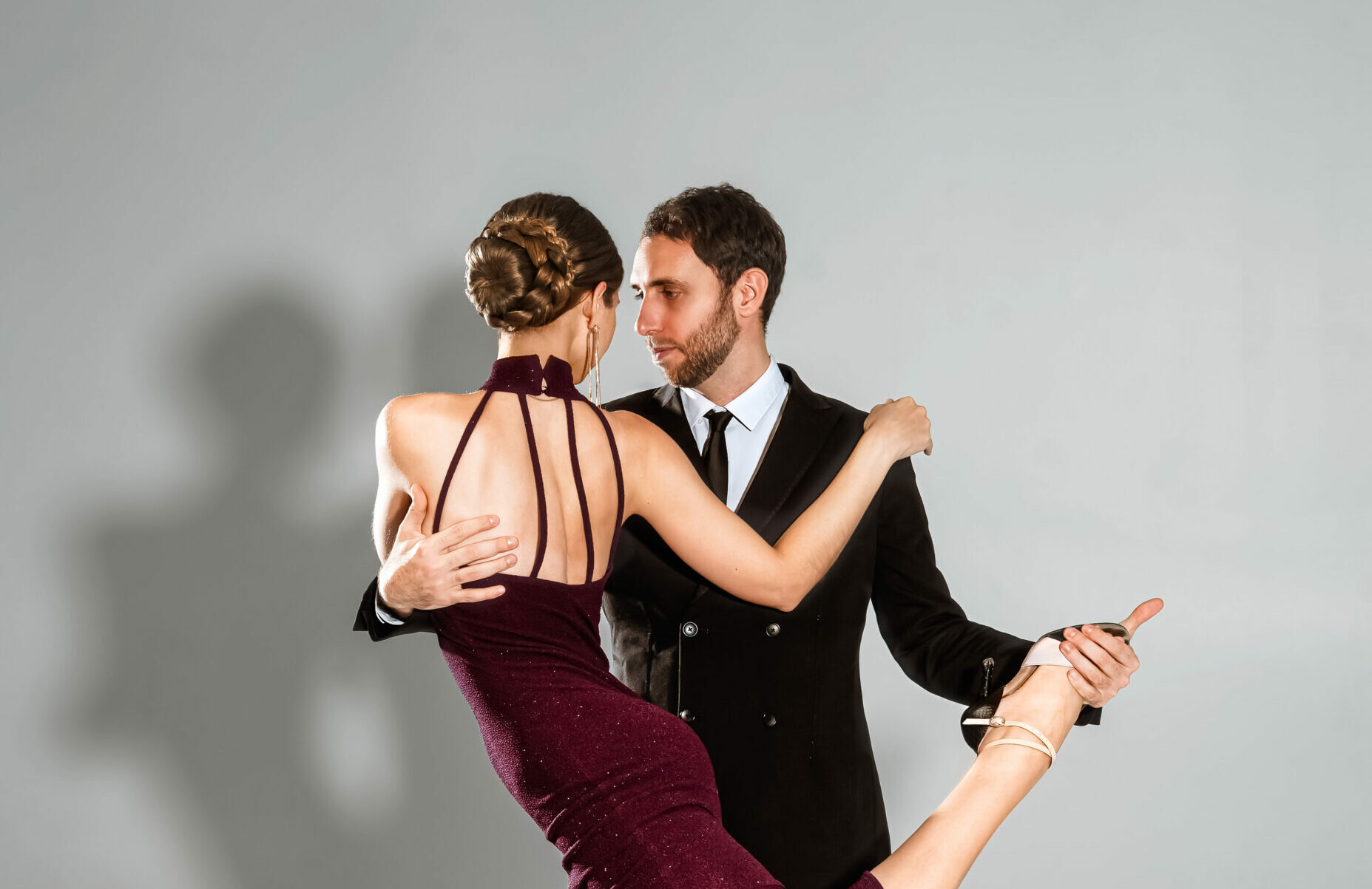 A full-body studio shot of two professional tango dancers. A woman in a shimmery plum dress with a ruffled hem balances on one leg while her partner, in a black suit, supports her lifted leg in a technical "gancho" maneuve